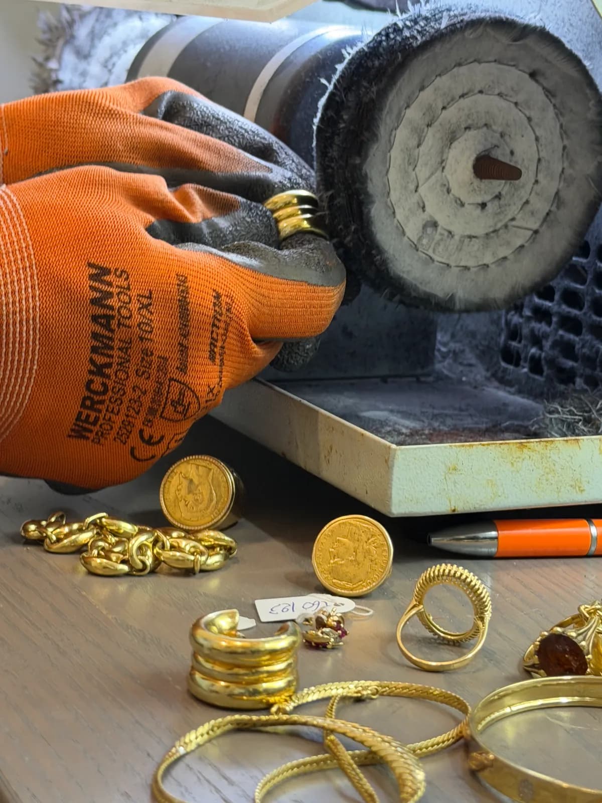 Mains gantées tenant une bague en or contre la brosse d'un touret de polissage, bijoux et pièces en or sur l'établi